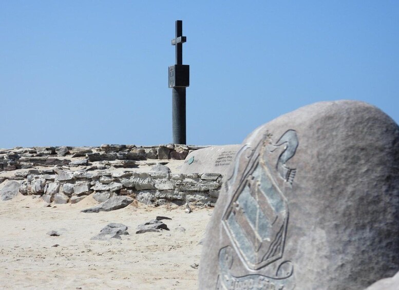 A stone monument with an engraved emblem, a stone wall, and a cross on a pole in the background. 2