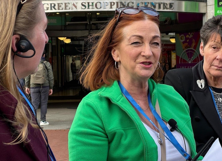 A person in a green jacket with a microphone, standing in front of a shopping center. 1