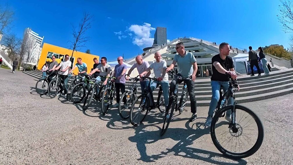 A group of cyclists on a paved path with a modern building in the background. 1