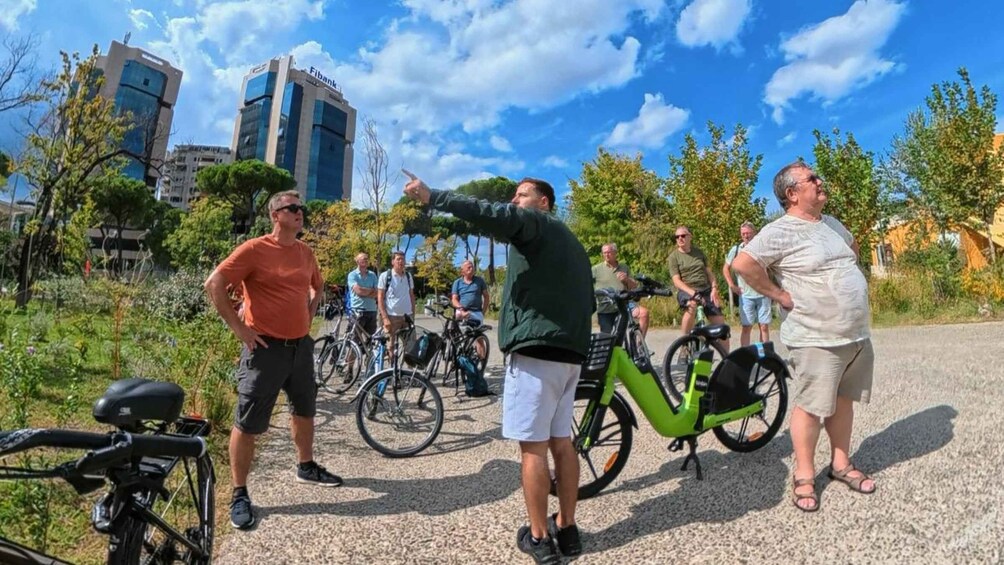 A group of people standing outdoors with bicycles, a building labeled 'Fibank' in the background, and a clear sky. 5