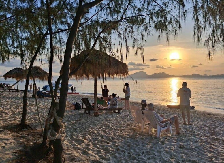 A beach with people relaxing under trees and umbrellas during sunset. 3