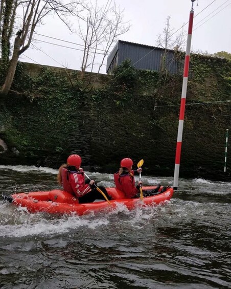 Two people in a red raft navigating a river with rapids, surrounded by trees and a building. 2