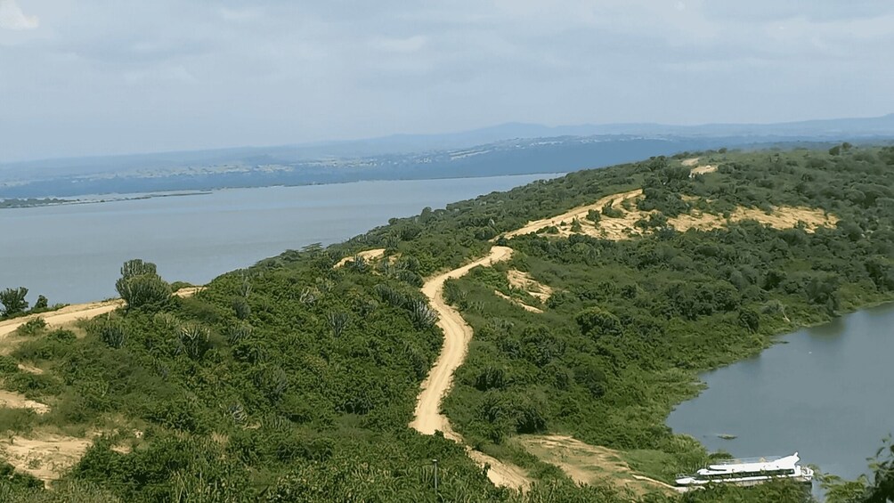 A winding dirt path through a lush green landscape beside a calm body of water, with distant mountains under a cloudy sky. 2