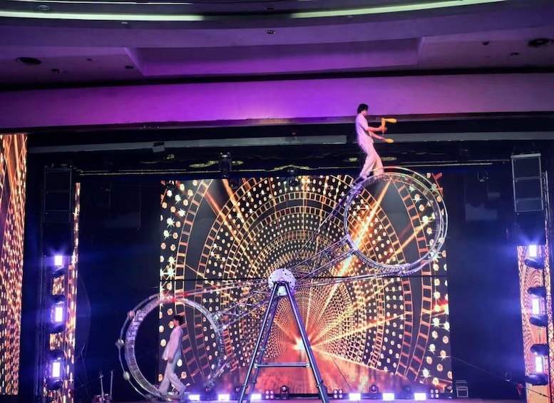 A circus performer on a unicycle balancing on a pole, with a large, illuminated circular structure in the background. 3