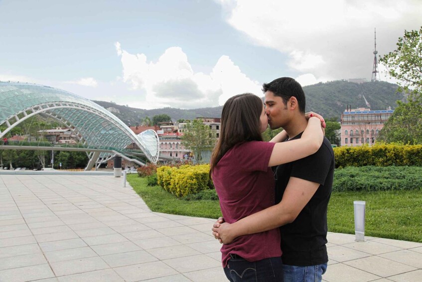 A couple embracing in a public square with a modern glass bridge and buildings in the background. 3