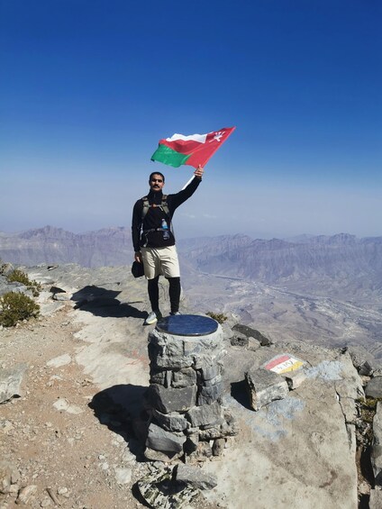 A person standing on a rocky outcrop holding a flag, with a mountainous landscape in the background. 5