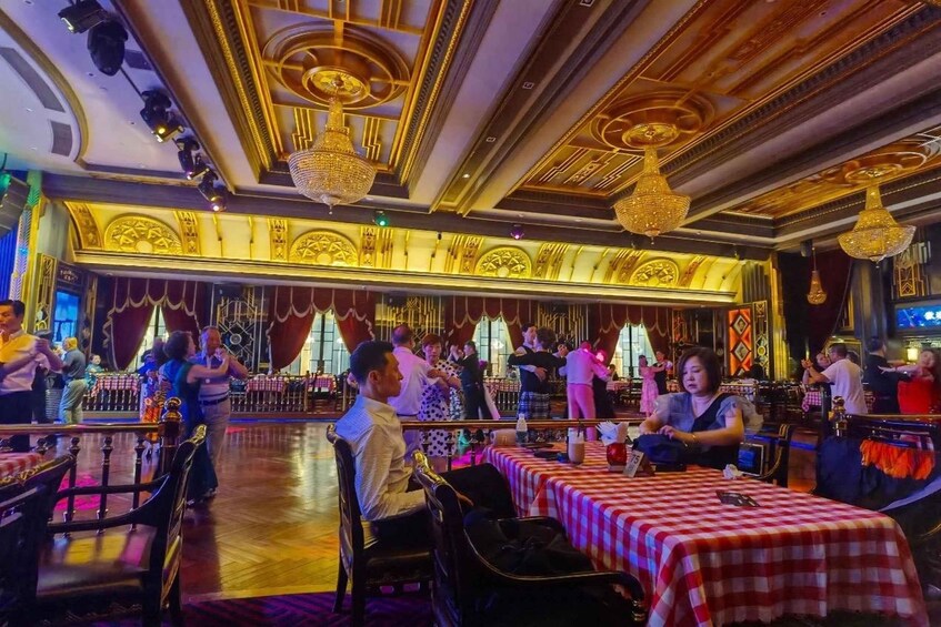 A ballroom with ornate ceiling and chandeliers, tables with checkered tablecloths, and people dancing. 2