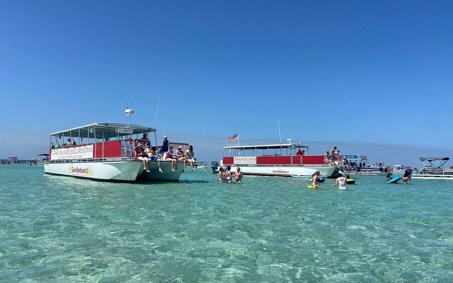 Two boats with people on them, one with a flag, and several people on the shore with surfboards. 2