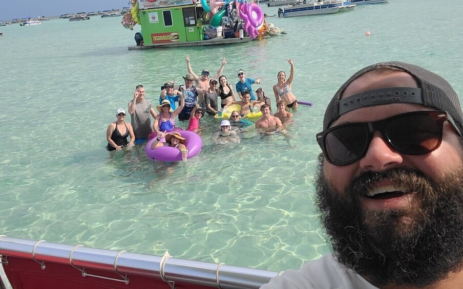 A group of people in a clear blue sea, some on a boat and others in the water, with a green boat in the background. 1