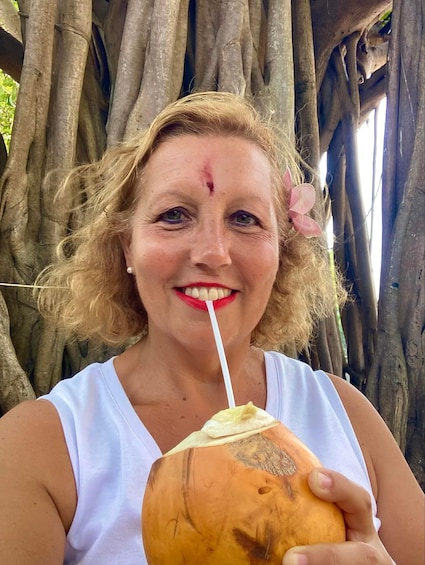 A person holding a coconut with a straw, standing in front of a tree with thick roots. 4