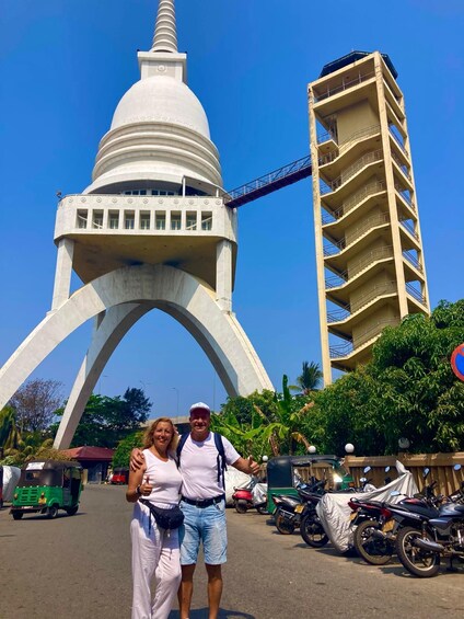 A couple posing in front of a large white monument with a bridge, surrounded by parked motorcycles and greenery. 3