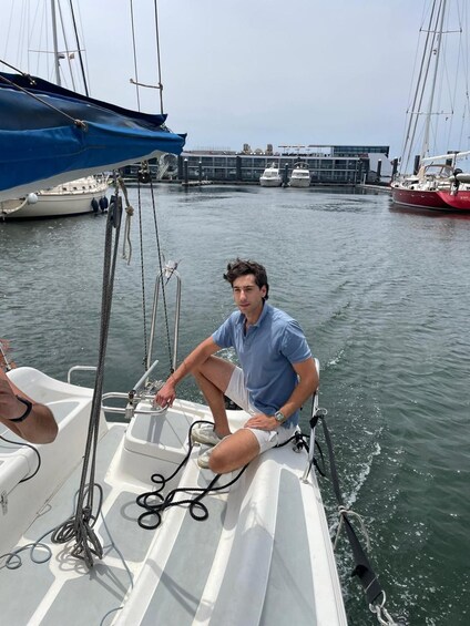 A person on a sailboat with ropes and a blue tarp, other boats in the background, and a building across the water. 3