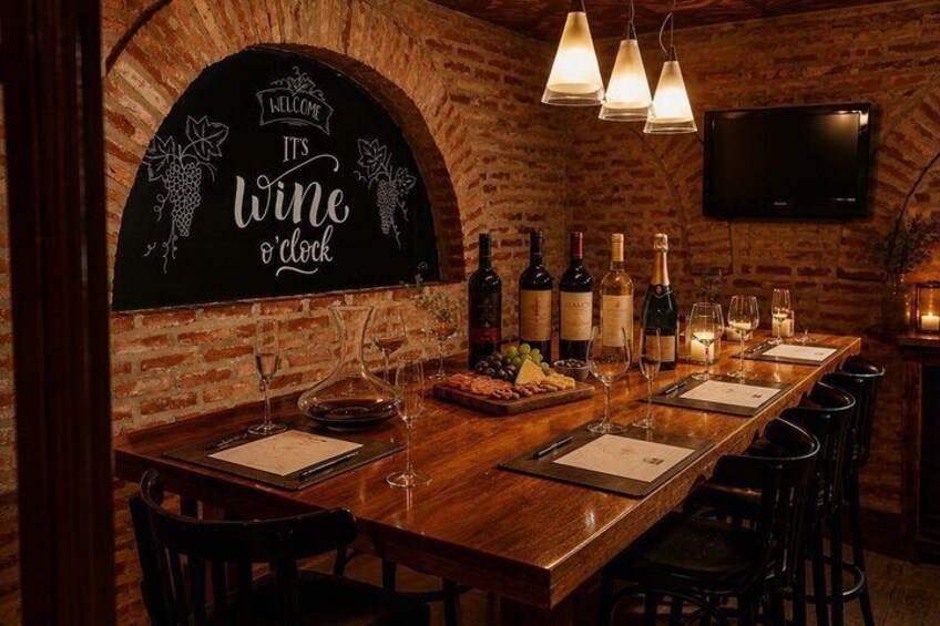 A brick-walled bar with a 'Welcome It's Wine o'Clock' sign, bottles, glasses, and a TV mounted on the wall. 2
