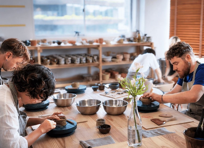 Un atelier de poterie où l’on voit des personnes travailler sur des pots en argile, divers outils de poterie et, au fond, des étagères remplies de pièces finies. 4