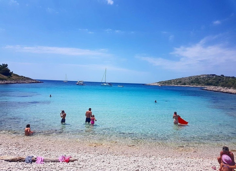 A pebbly beach with clear turquoise water, people swimming and sunbathing, and sailboats in the distance. 4