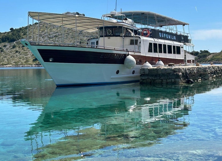 A boat named 'EUFEMIJIA' docked at a calm harbor with clear water reflecting the boat and surrounding landscape. 1