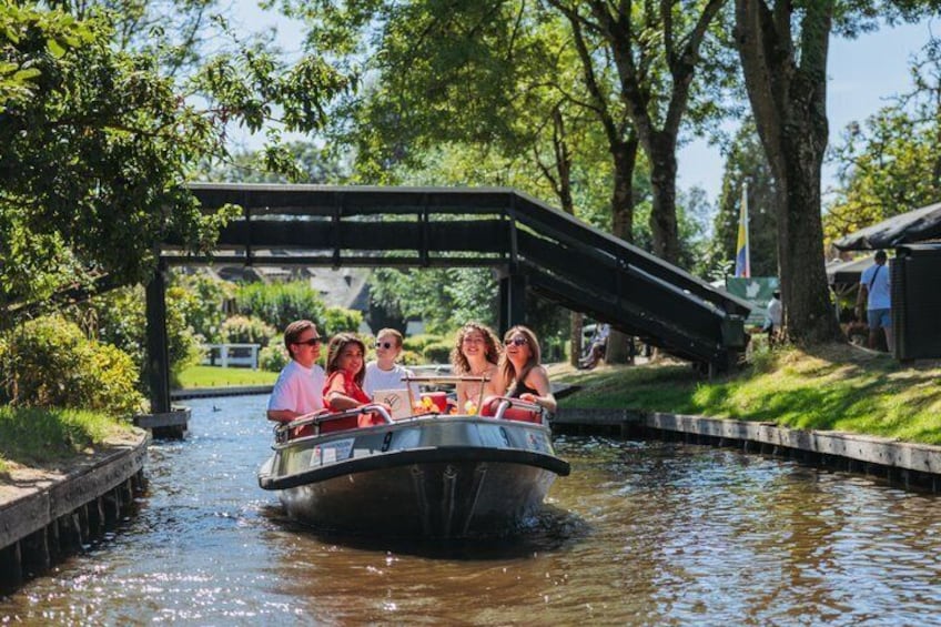 A group of people on a narrowboat on a canal, with a bridge overhead and greenery around. 1