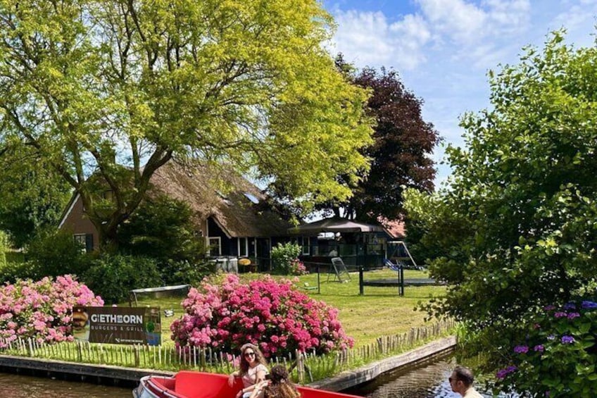 A riverside restaurant with a thatched roof, surrounded by blooming pink flowers and lush greenery. 1