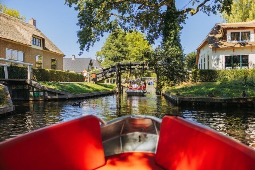 A narrow canal with a red boat in the foreground, a wooden bridge, and houses on either side. 3