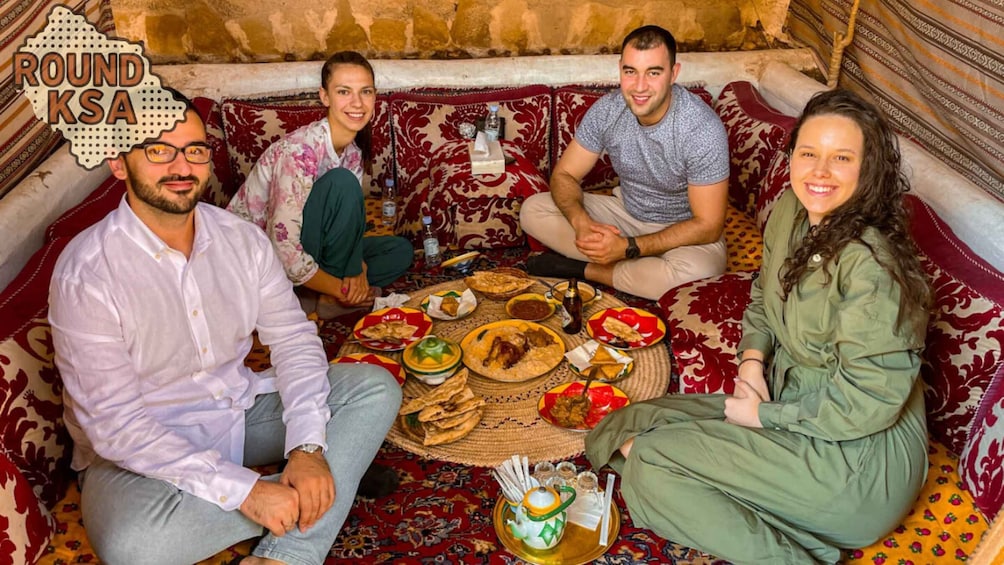 A group of people sitting on a patterned sofa around a table with various dishes and a teapot. 1