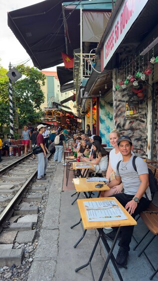 A bustling street market with people sitting at wooden tables, a Vietnamese flag, and a sign for a 56 Dong restaurant. 5
