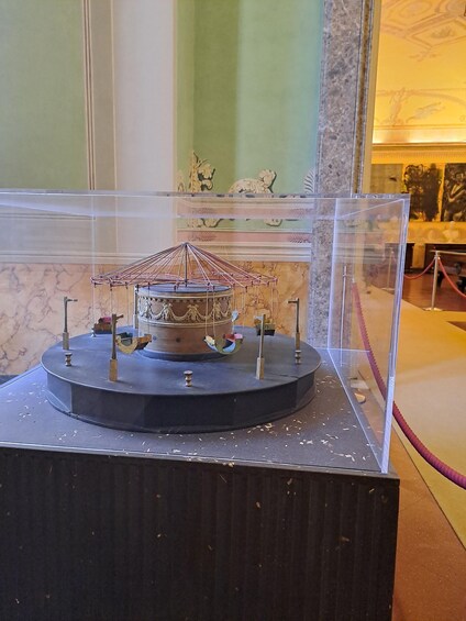 A historical carousel model displayed in a glass case, with a marble floor and classical wall decorations in the background. 2
