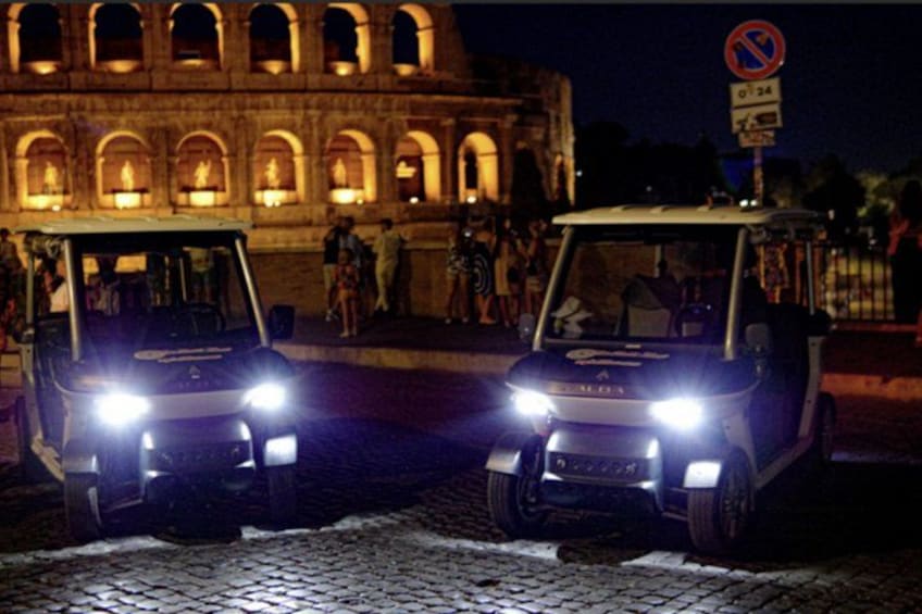 Two golf carts with headlights on in front of a historic building with arches. 2