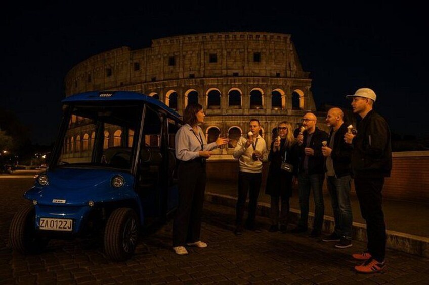 A group of people standing in front of the Colosseum at night with a blue car parked nearby. 3