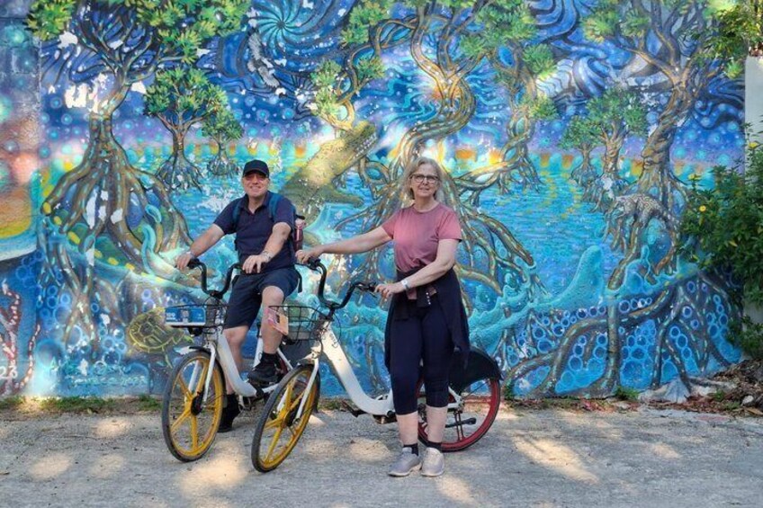 Two people on bicycles in front of a mural with tree and ocean designs. 1