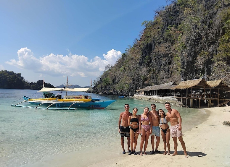 A group of people on a beach with a boat and a thatched-roof structure in the background. 1