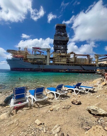 A large cargo ship docked at a beach with clear turquoise water and blue lounge chairs on the sand. 4