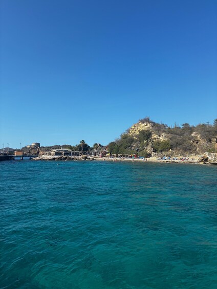 A coastal landscape with clear blue waters, a rocky hill with sparse vegetation, and a distant building on the shore. 1