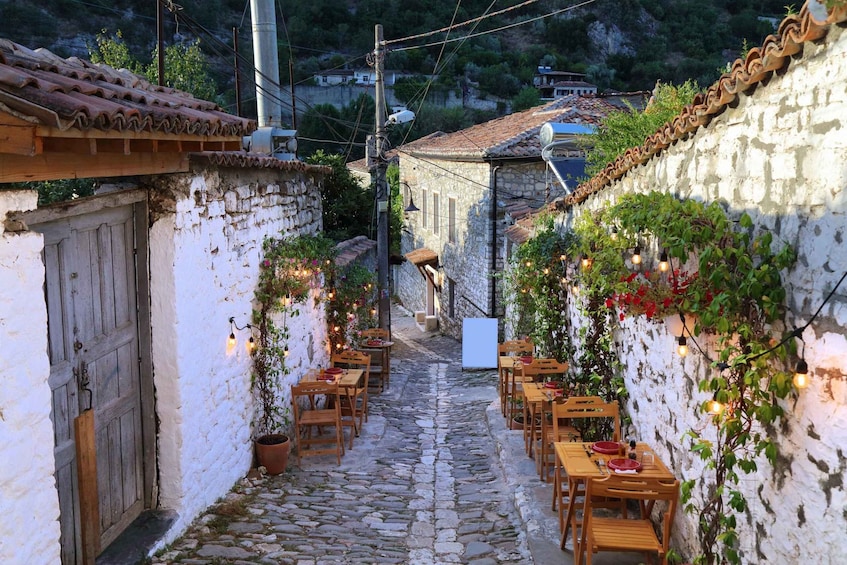 Un estrecho callejón adoquinado con edificios de piedra, mesas y sillas de madera dispuestas para comedor al aire libre, rodeado de vegetación e iluminación decorativa. 5