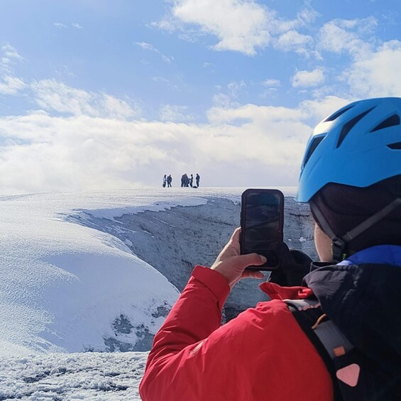 A person in a red jacket and blue helmet taking a photo of a group of people on a snowy mountain. 4