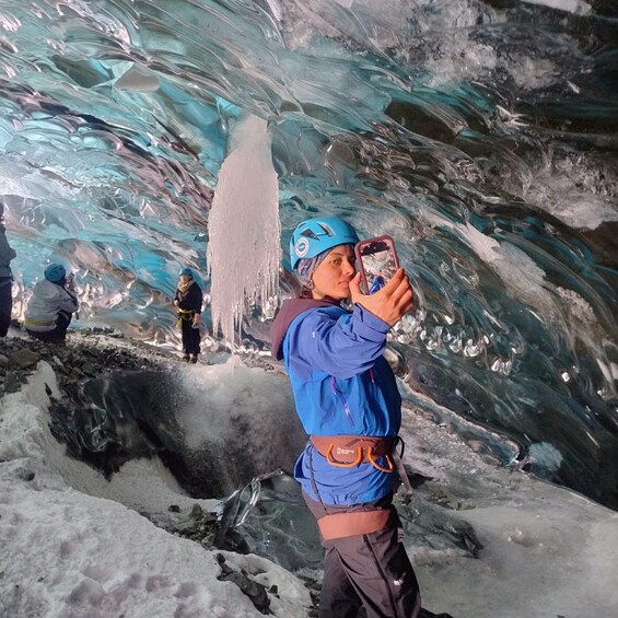 A person in a blue jacket and helmet taking a photo inside an ice cave with icicles hanging from the ceiling. 3