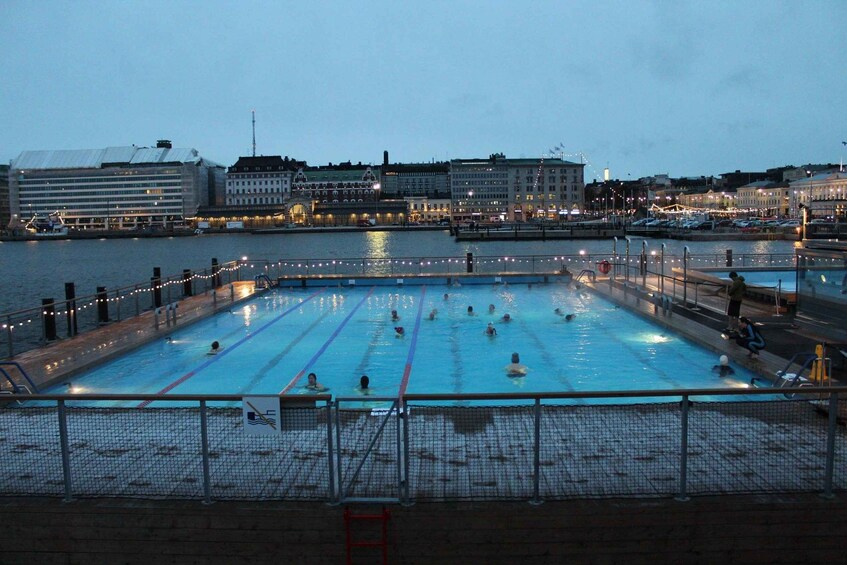 A swimming pool with people in the water, surrounded by a cityscape with buildings and a waterfront. 5