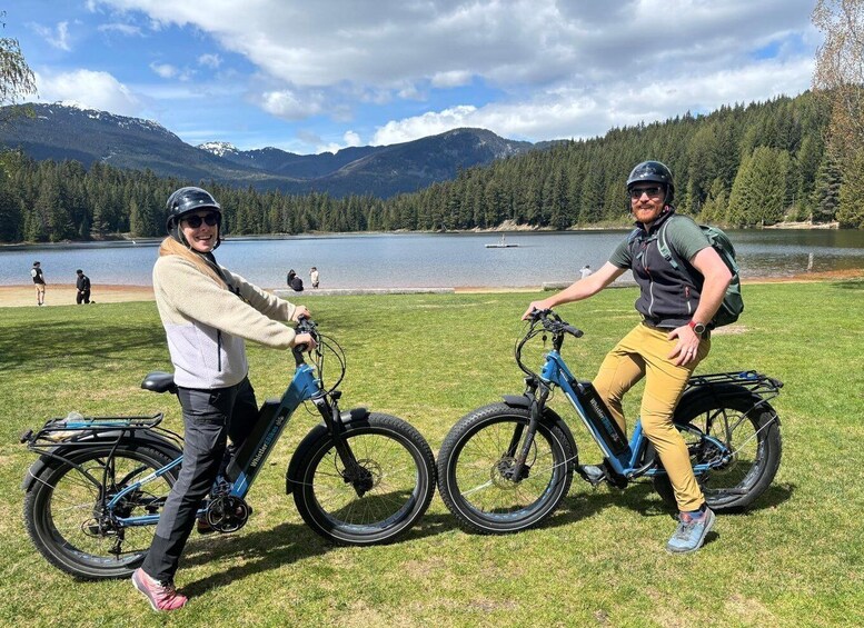 Two people on electric bikes by a lake with mountains in the background. 3