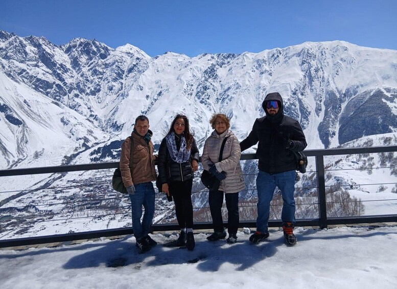 A group of people standing on a snowy ledge with a mountain range in the background. 1