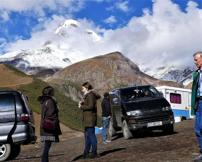 A group of people standing near a black vehicle with a camper trailer, with a snow-capped mountain in the background. 3