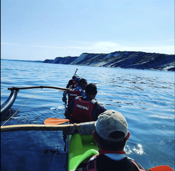 A group of people on a boat with 'OKI BA' life jackets, paddling near a coastline with cliffs. 2