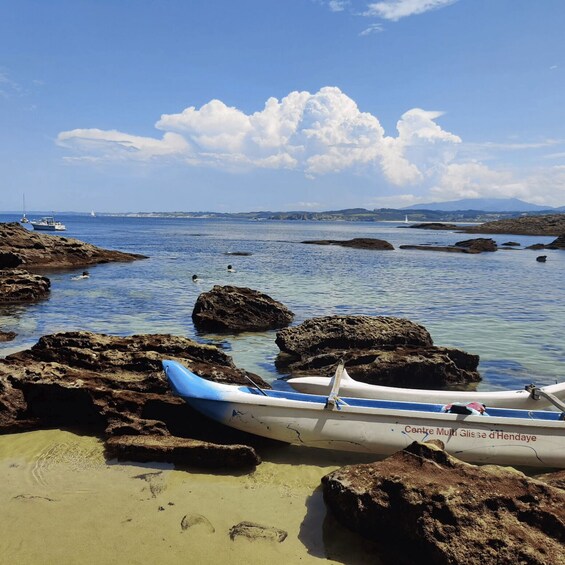 A rocky shoreline with two canoes, clear blue water, and distant boats. 1