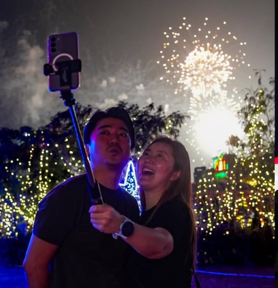A person holding a selfie stick with a smartphone, standing in front of a fireworks display. 2