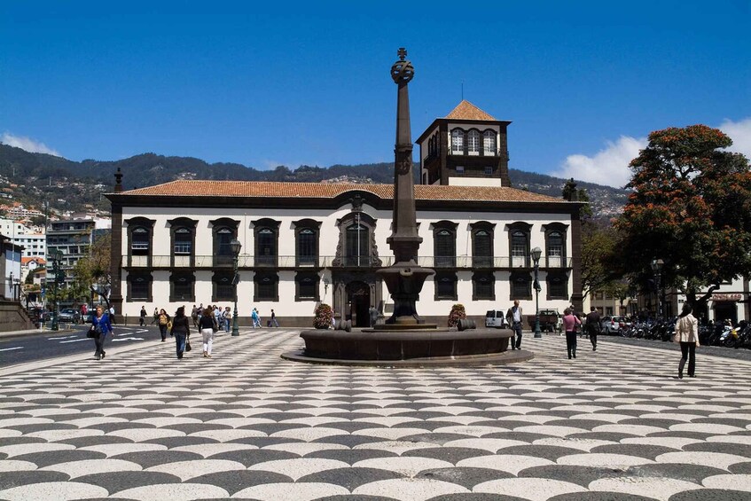 A plaza with a fountain, a building with a tower, and people walking. 5