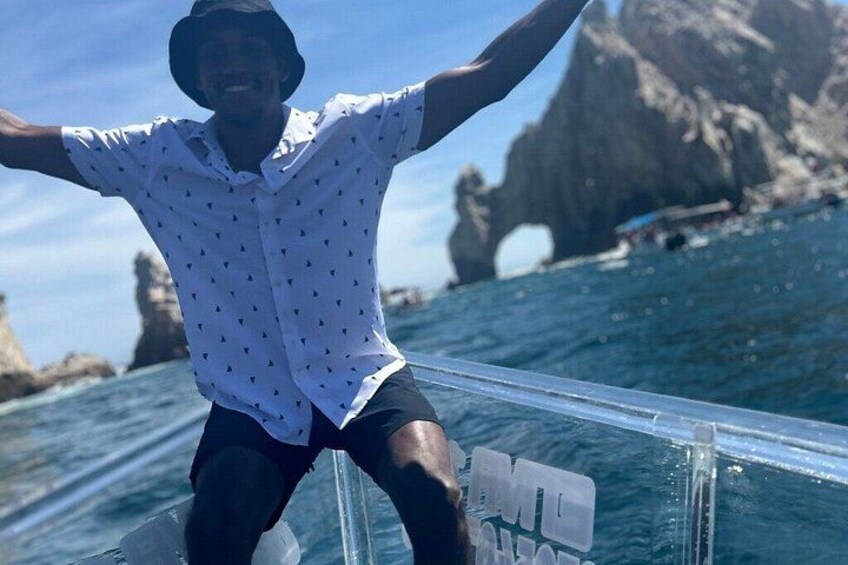 A person sitting on a transparent railing with arms outstretched, overlooking a body of water with a rocky coastline in the background. 4