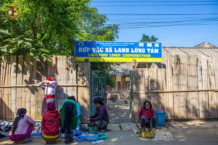 A group of women sitting and standing in front of a bamboo fence, with a signboard above them. 4