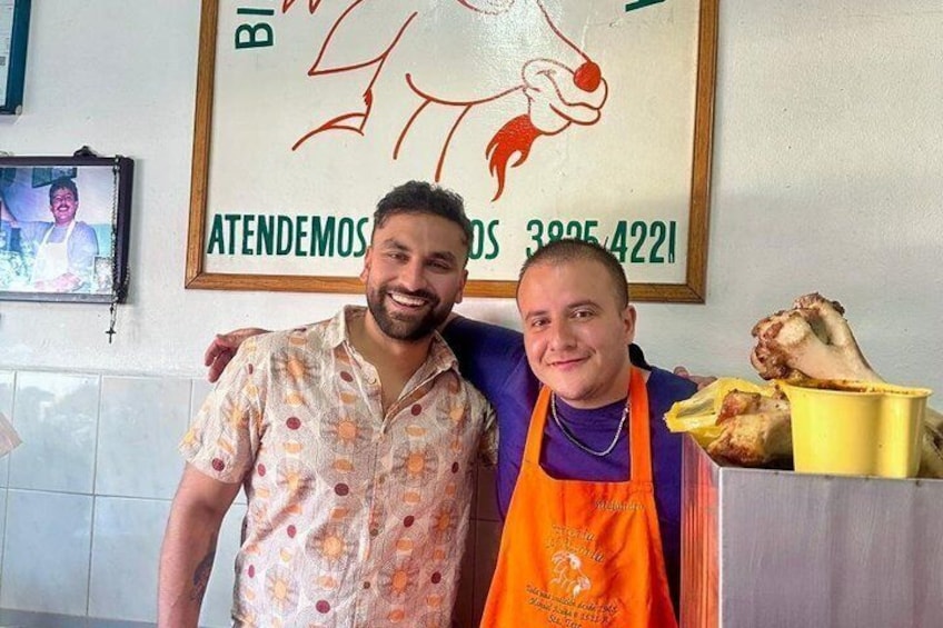 Two people in a kitchen, one wearing a patterned shirt and the other in an apron, with a framed poster and a yellow bucket on the counter. 5