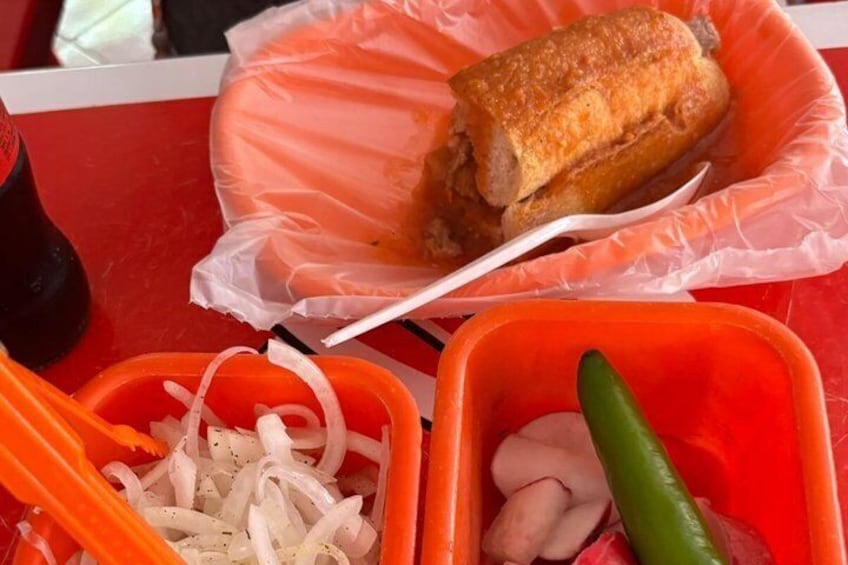 A sandwich in a plastic container, sliced onions, and a green chili pepper. 3
