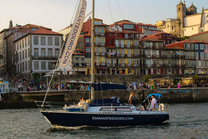 A sailboat with 'DIAMANTEDOURO.COM' on its side, docked near a waterfront with colorful buildings in the background. 2