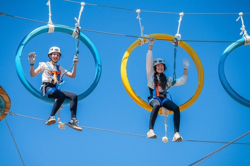 To personer på en zipline, iført hjelme og seler, med en klar blå himmel i baggrunden. 1