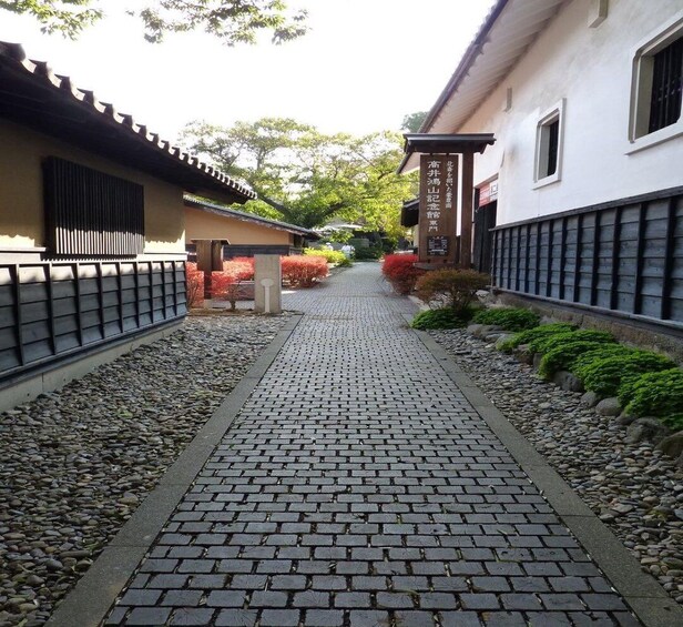 A traditional Japanese pathway lined with stone tiles, bordered by gravel and small shrubs, leading to a building with a signboard. 3